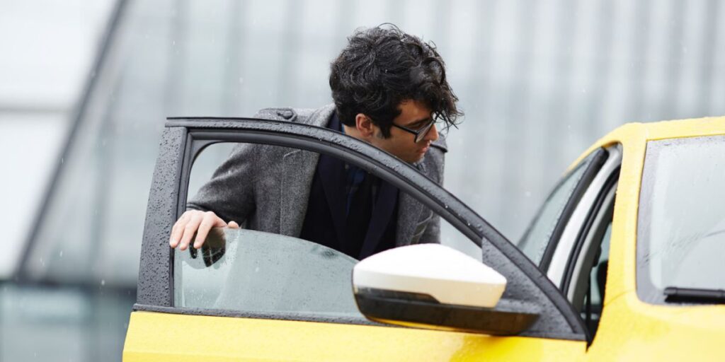 A man in a grey coat and glasses leans into a yellow car on a rainy day, with his hand on the open door's window.