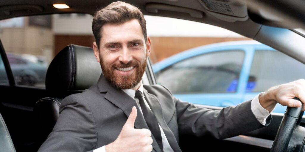 A smiling male driver in a suit and tie gives a thumbs up while sitting behind the steering wheel of a black car.
