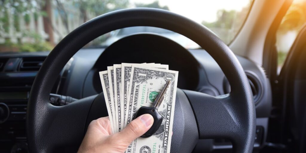 A person’s hand holds a car key and a fan of US dollar bills in front of a steering wheel inside a vehicle.