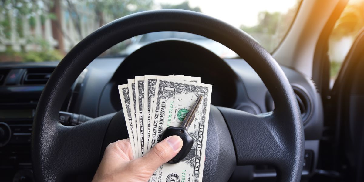 A person’s hand holds a car key and a fan of US dollar bills in front of a steering wheel inside a vehicle.