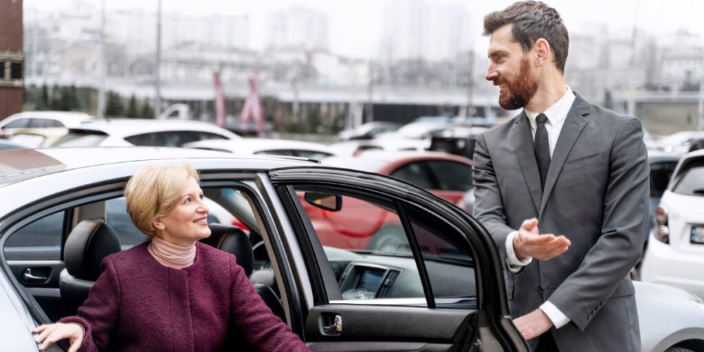 A professional male driver in a suit smiles while helping a woman in a purple coat exit a silver car in a parking lot.