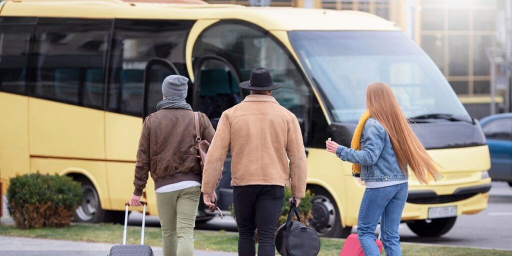 Three travelers with luggage walk toward a large yellow airport shuttle bus parked on a city street during the day.