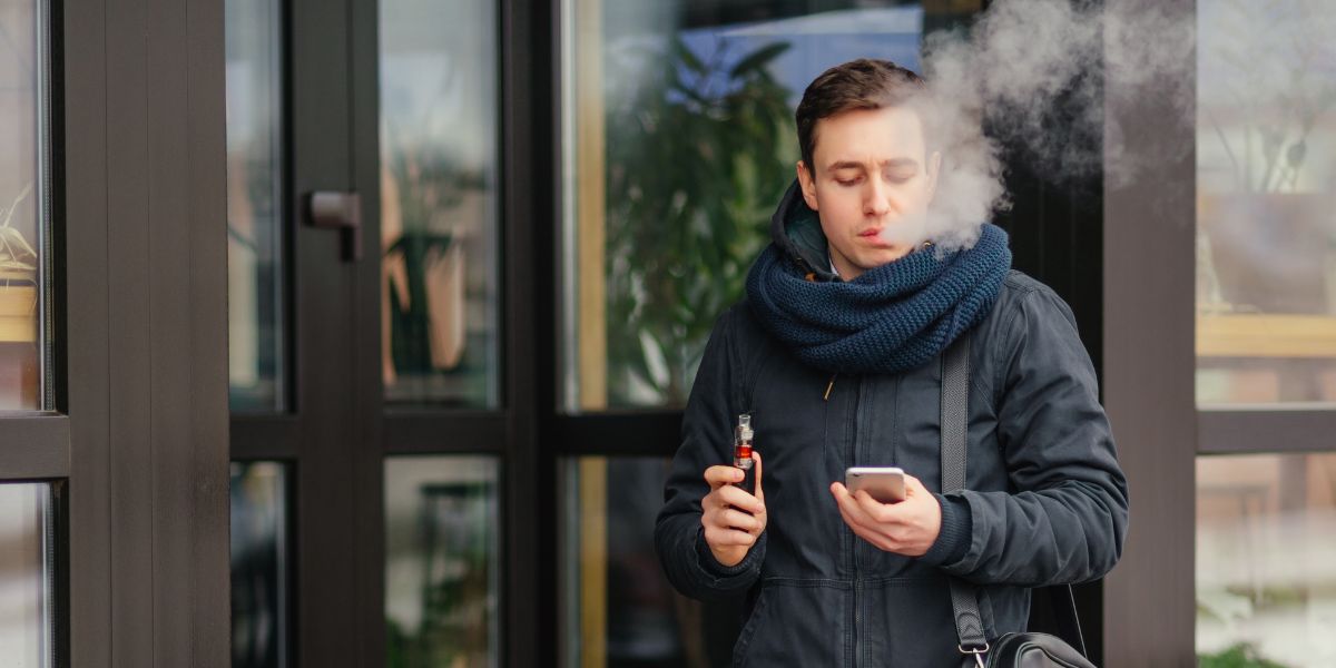 A man in a dark jacket and blue scarf uses a vape device while looking at his smartphone outside a building