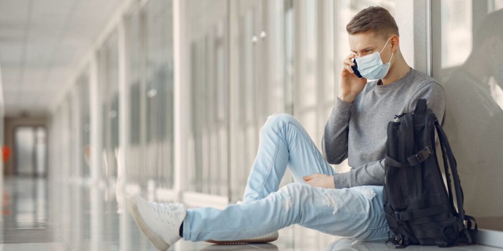 A young man wearing a face mask and casual clothes sits on a terminal floor while talking on his mobile phone.