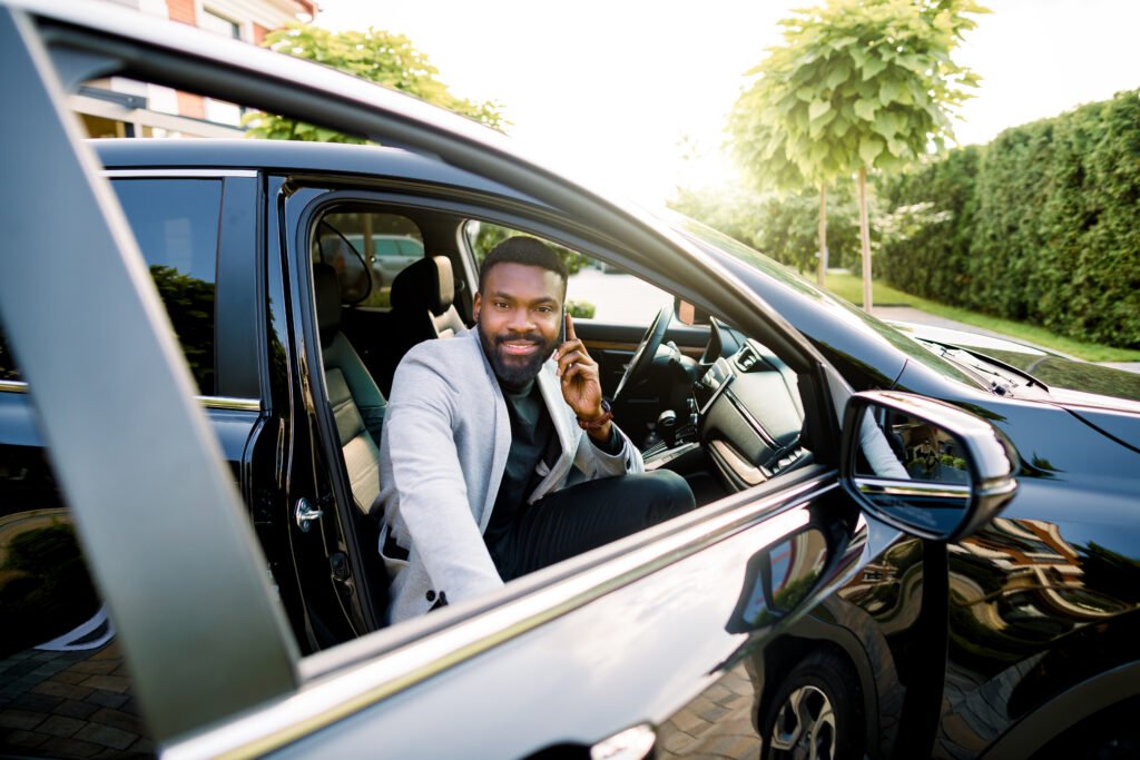 Happy smiling African american businessman in smart casual suit speaking on smartphone while getting in the black car on passenger seat.